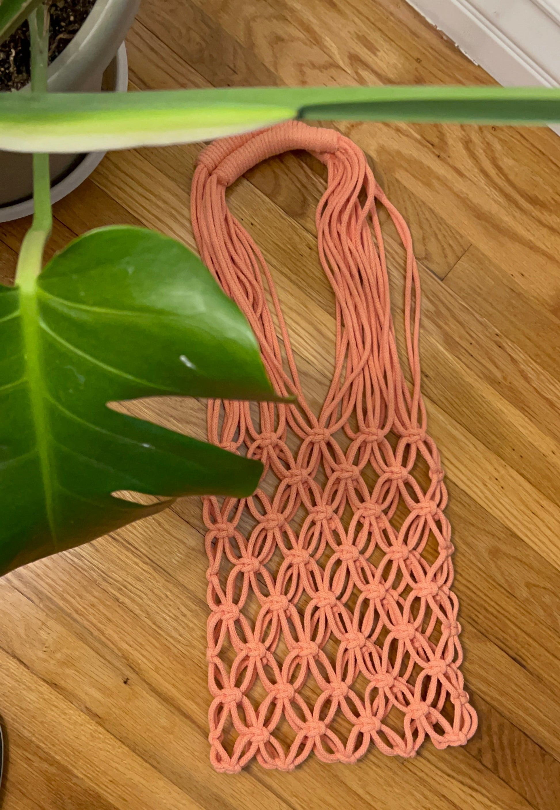 A peach-pink macrame tote bag with a geometric woven pattern, laid flat on a light brown wooden floor next to a green plant leaf.