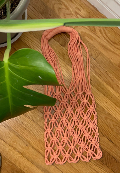 A peach-pink macrame tote bag with a geometric woven pattern, laid flat on a light brown wooden floor next to a green plant leaf.