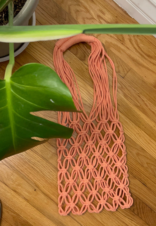 A peach-pink macrame tote bag with a geometric woven pattern, laid flat on a light brown wooden floor next to a green plant leaf.